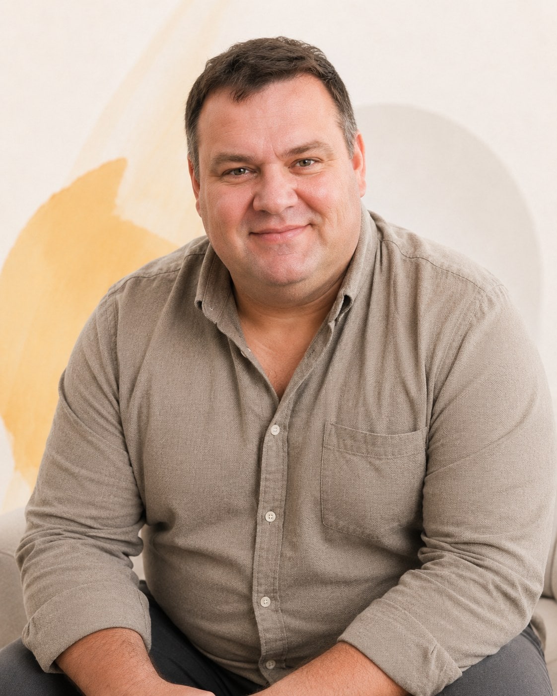 A heavyset man in a taupe button-down, leaning forward slightly with a small, easy smile.