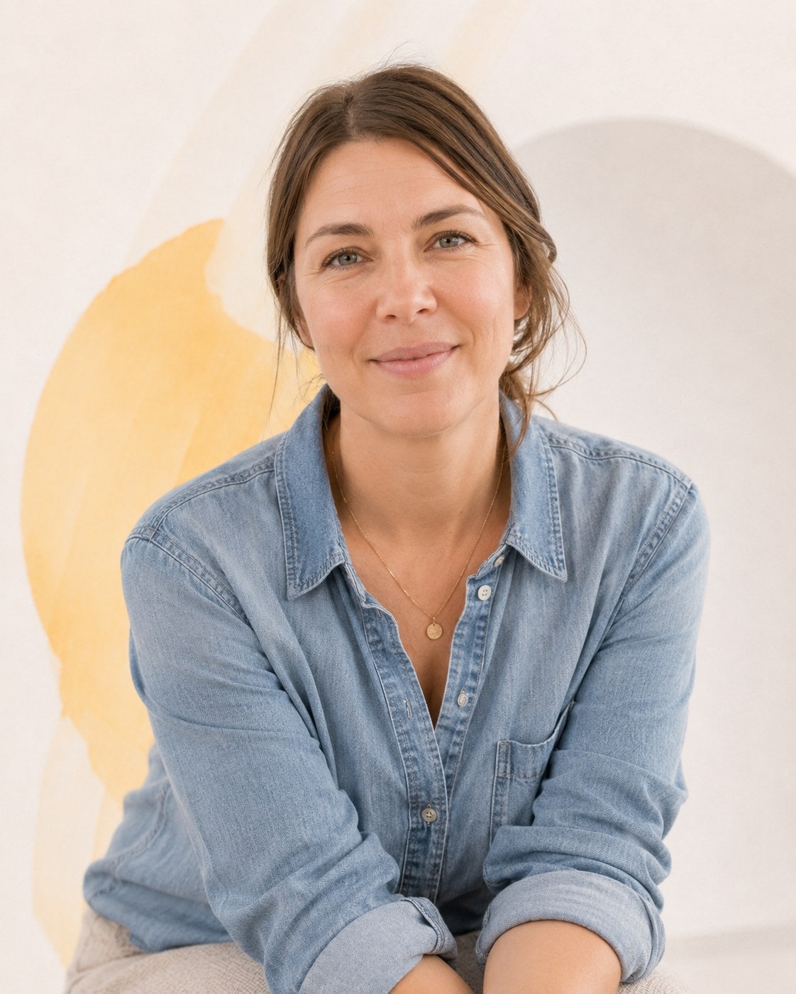 A woman in a denim shirt with her hair pinned up, forearms on her knees, looking directly ahead.