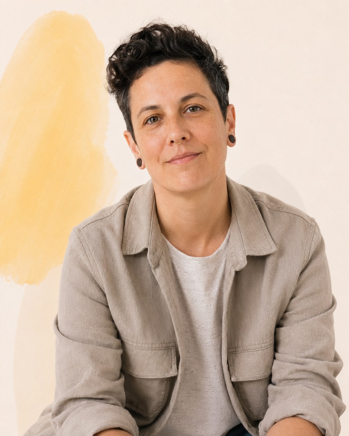 A person with short dark curly hair and ear piercings in a taupe overshirt, meeting the camera evenly.