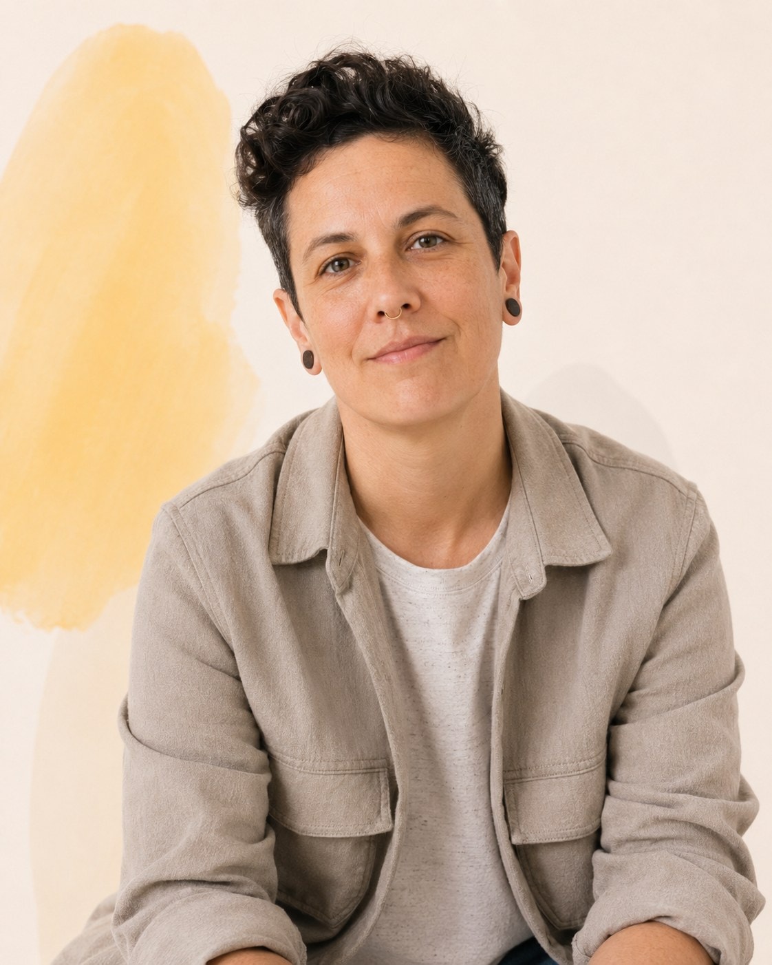 A person with short dark curly hair and ear piercings in a taupe overshirt, meeting the camera evenly.